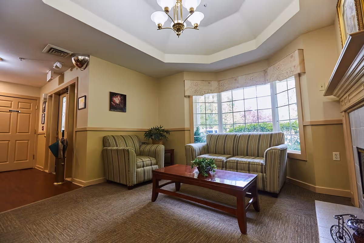A cozy senior living facility common area with a striped sofa and matching armchair around a wooden coffee table with a small plant. Large windows with beige valances let in natural light, and a fireplace is visible on the right. The room has a beige and light green color scheme with a chandelier hanging from a tray ceiling.
