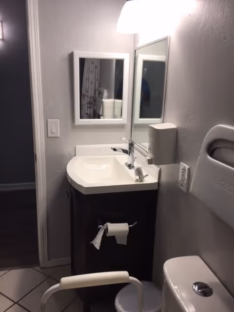 Small bathroom with a white sink and dark brown cabinet, a mirror above the sink, a wall-mounted soap dispenser, a toilet with a grab bar, and a toilet paper holder with a roll of toilet paper. The walls are light gray and the floor is tiled.