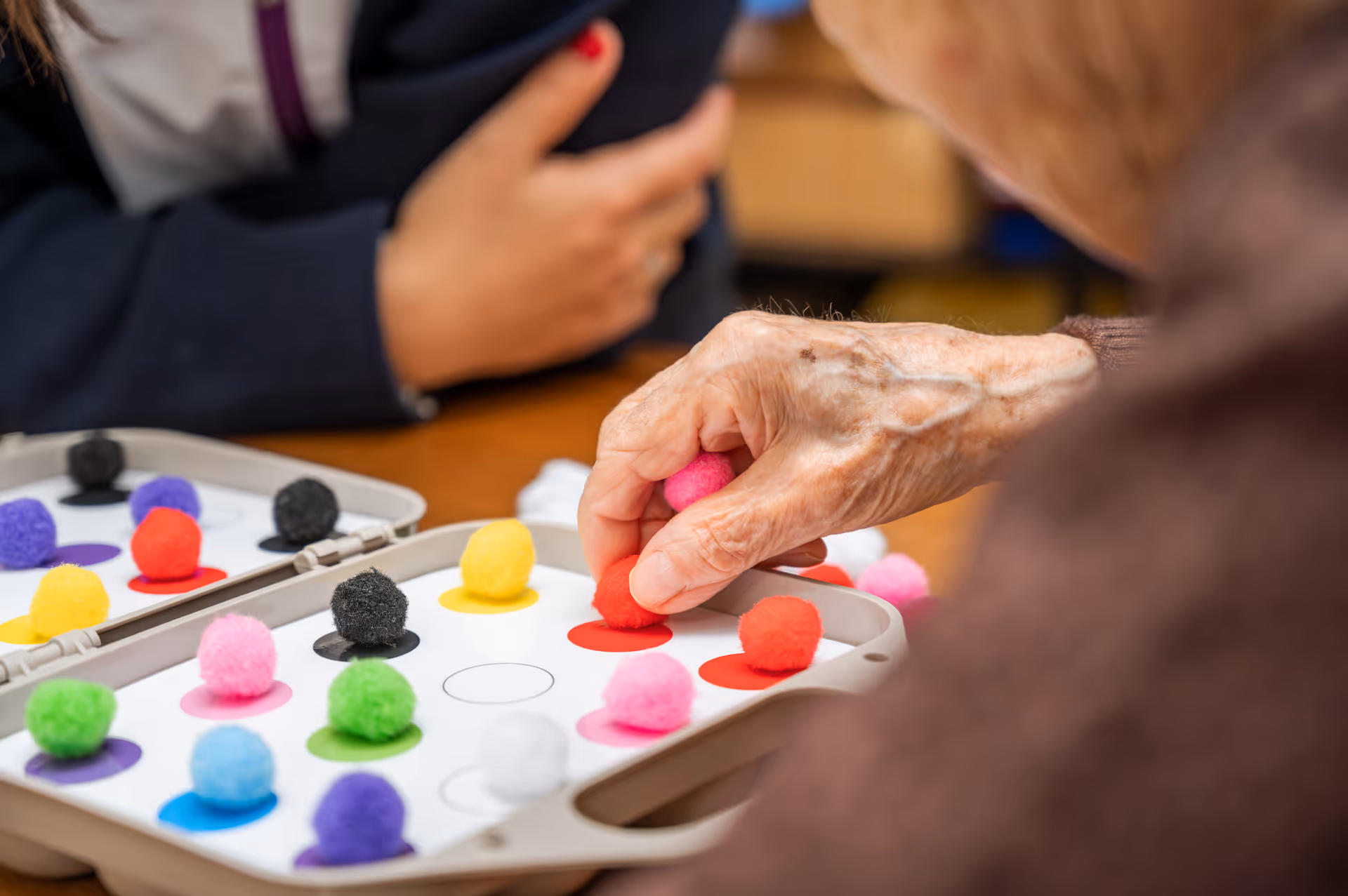 Close-up of an elderly person's hand placing a red pom-pom on a game board with colorful pom-poms, with another person in the background.