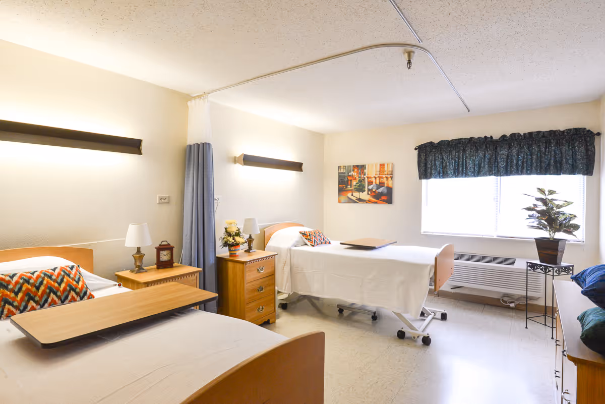 Shared senior care bedroom with two hospital-style beds, bedside tables, overhead lights, a curtained divider and a window with a potted plant.