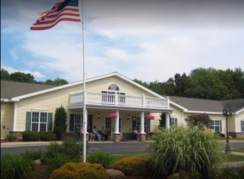 Exterior view of a single-story yellow senior living facility building with white trim and a balcony above the entrance. There is an American flag on a flagpole in front of the building, surrounded by landscaped bushes and plants. Several people are sitting on the porch under the covered entrance.