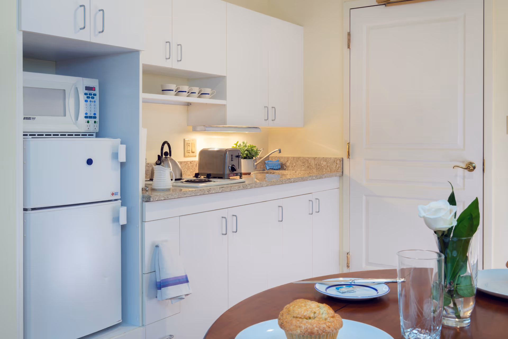 A small kitchen area with white cabinets, a microwave, a refrigerator, a toaster, a kettle, and a sink. In the foreground, there is a wooden table set with plates, a glass, and a muffin, along with a vase holding a white rose.
