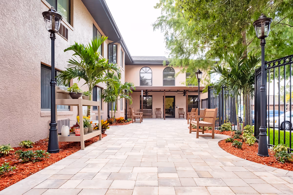 Outdoor courtyard area of a senior living facility with a paved walkway, benches, potted plants, palm trees, and black lamp posts. The building has beige walls and multiple windows, with a covered seating area at the far end.