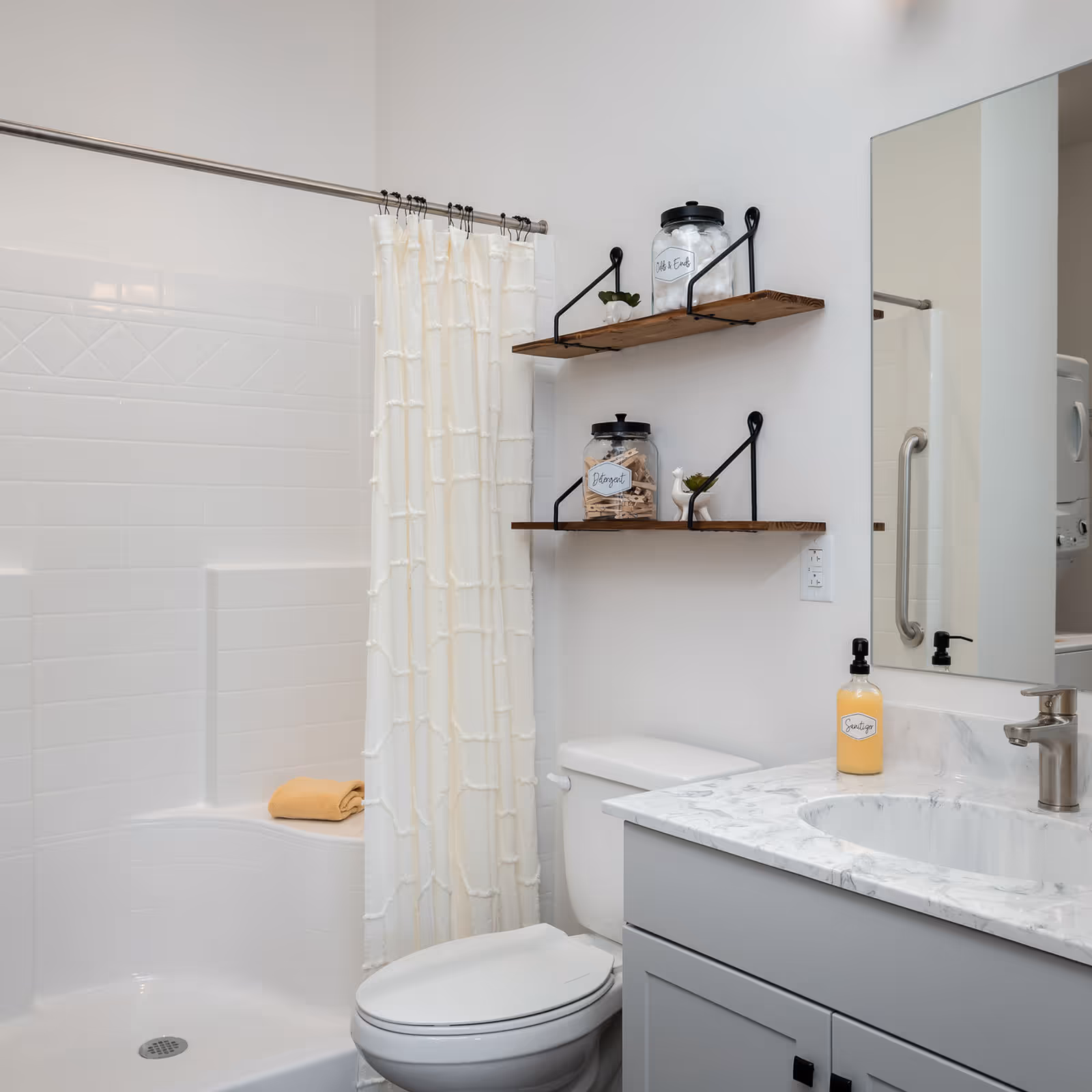 A clean and modern bathroom featuring a white tiled shower with a cream-colored textured shower curtain, a white toilet, and a gray vanity with a marble countertop and an oval sink. Above the toilet, there are two wooden shelves holding labeled jars and small decorative plants. A large mirror is mounted above the vanity, and a bottle of sanitizer is placed on the countertop.