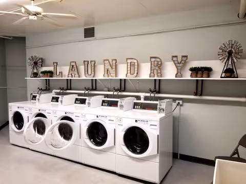 A bright laundry room with five front-loading washing machines beneath a shelf displaying marquee letters that spell "LAUNDRY" and decorative plants.