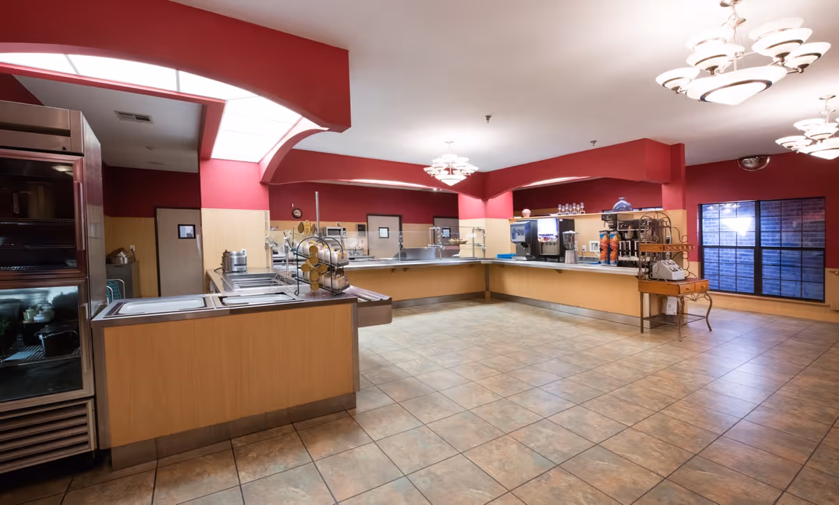 A spacious senior living facility kitchen area with a long counter for serving food, beverage dispensers, and a refrigerated display case. The walls are painted red with beige paneling, and the floor is tiled. Ceiling lights illuminate the room.