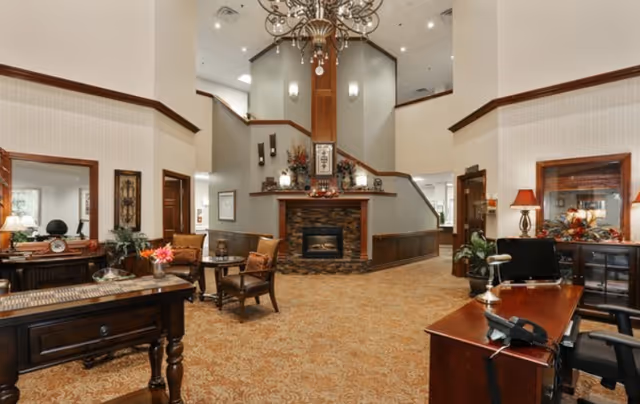 Spacious and warmly decorated senior living facility common area with a central stone fireplace, high ceilings, and a chandelier. The room features comfortable seating with armchairs and tables, wooden desks with lamps and office equipment, and decorative plants and artwork. The carpet has a patterned design, and the walls are painted in neutral tones with wood trim accents.