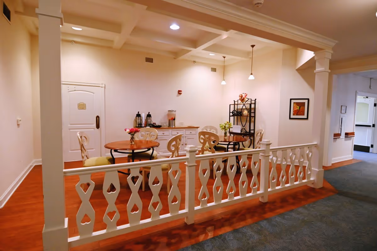 A cozy seating area in a senior living facility with two round wooden tables, each surrounded by decorative chairs. The area is enclosed by a white wooden railing with heart-shaped cutouts. There is a white cabinet against the wall with coffee and beverage dispensers on top, and a black metal shelf with decorative items. The walls are light-colored, and the floor is a combination of wood and carpet. Ceiling lights and pendant lamps illuminate the space.