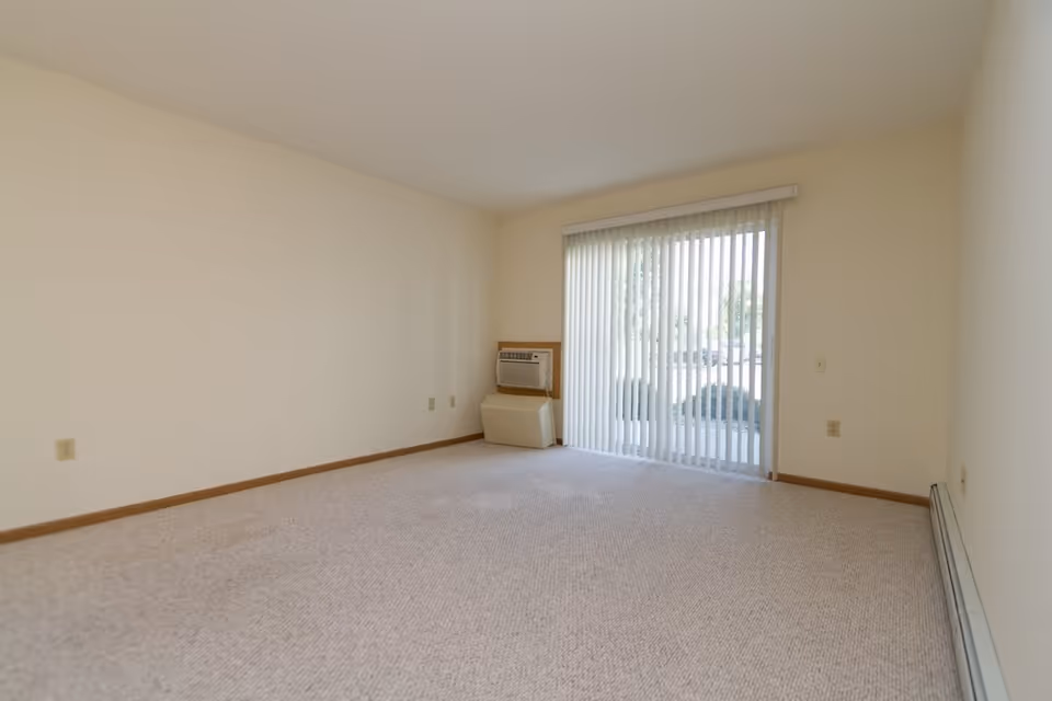 Empty carpeted living room with a sliding glass door and vertical blinds, a wall air conditioner, and baseboard heating.
