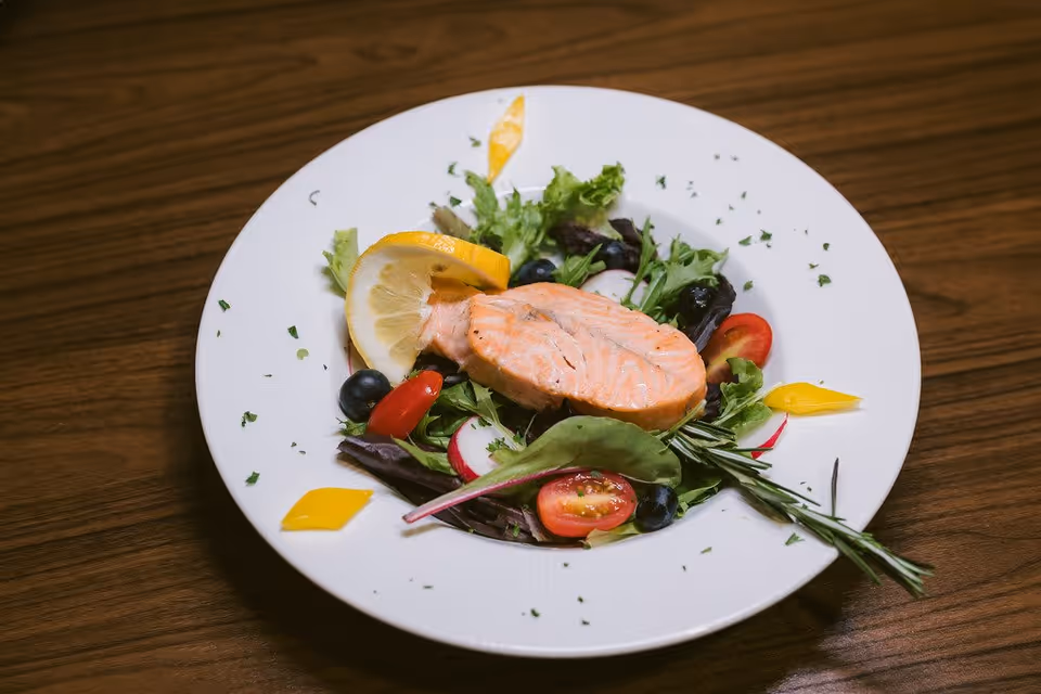 A white plate with a piece of cooked salmon placed on a bed of mixed greens, cherry tomatoes, radish slices, black olives, and garnished with a lemon wedge and herbs, set on a wooden table.