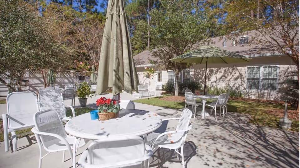 Sunny outdoor patio with white tables and chairs, green umbrellas, potted plants, and the facility building in the background.