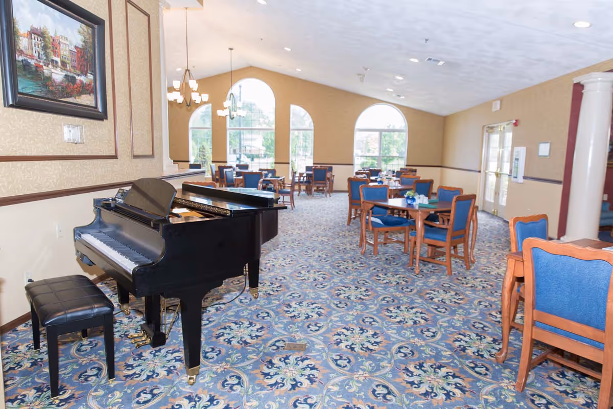 Spacious senior living dining/activity room with a black grand piano in the foreground and multiple tables and chairs under arched windows.