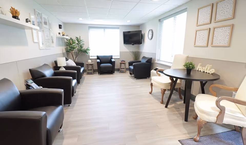 Bright communal lounge with multiple armchairs arranged along the walls, a small round table with chairs, a potted plant and a wall-mounted TV.