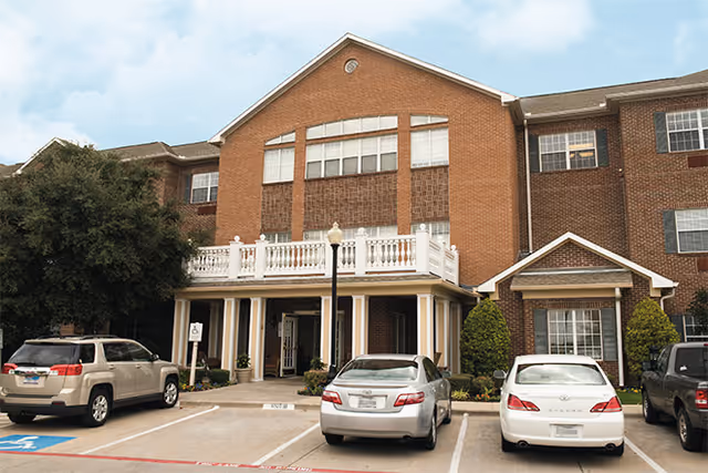 Brick three-story senior living facility front entrance with a balcony and cars parked in the lot