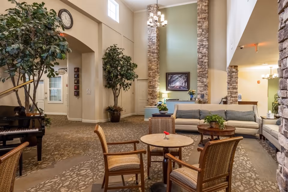 A spacious and well-lit living room area in a senior living facility featuring a beige sofa with striped cushions, a round wooden coffee table with a plant, and a small round table with four wooden chairs. The room has tall stone pillars, a large wall clock, indoor potted trees, a grand piano, and a chandelier hanging from the high ceiling. The walls are painted in neutral tones with a framed picture hanging above a small counter.