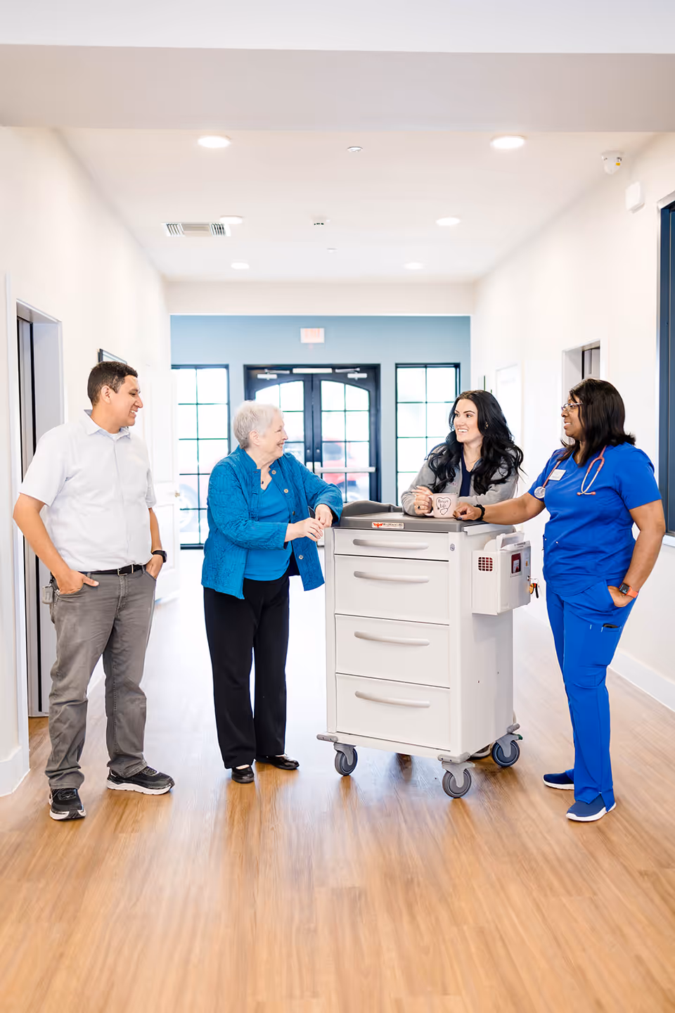Four people standing in a bright hallway with wooden flooring. An elderly woman in a blue jacket is talking with a man in a white shirt and gray pants, a woman with long dark hair holding a mug, and a nurse in blue scrubs with a stethoscope around her neck. They are gathered around a white medical cart with drawers on wheels.