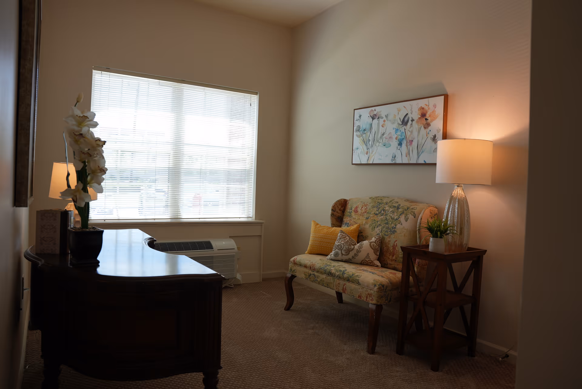 Cozy seating area with a floral loveseat, side table and lamp opposite a wooden desk by a window with blinds.