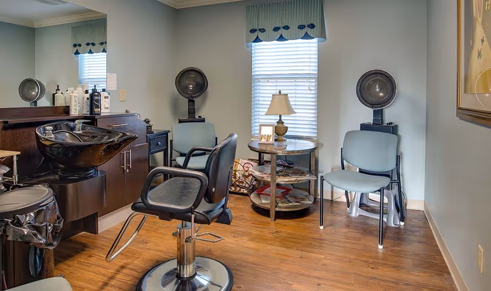 Interior of a salon area with a black salon chair in front of a hair washing station, two blue chairs with hair dryers behind them, a round table with a lamp and magazines, and a window with blinds and a blue valance.