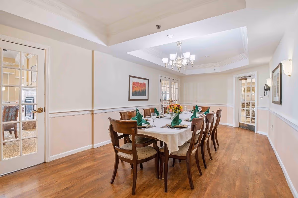 A dining room with a long rectangular table covered with a white tablecloth, set with plates, glasses, and green folded napkins. There are eight wooden chairs around the table. The room has wooden flooring, light-colored walls with a chair rail, a chandelier hanging from a tray ceiling, and framed artwork on the walls. Two glass-paneled doors lead to adjacent rooms.