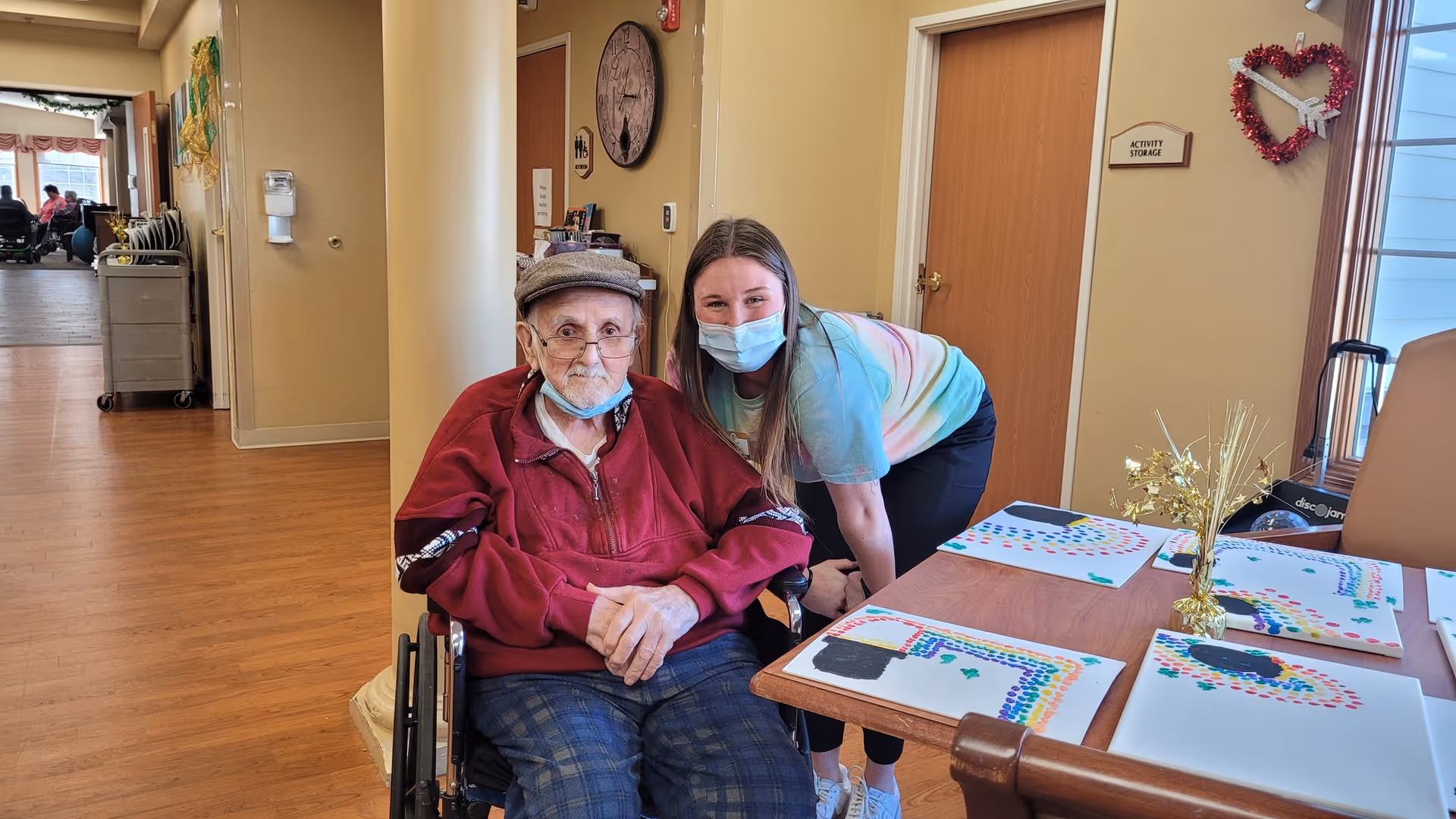 An elderly man in a wheelchair wearing a red jacket and a flat cap sits next to a young woman wearing a face mask and a tie-dye shirt. They are in a hallway or common area of a facility with wooden floors and beige walls. On the table in front of them are colorful artwork pieces and a small gold decorative centerpiece. A door labeled 'Activity Storage' and a heart-shaped decoration are visible on the wall behind them.