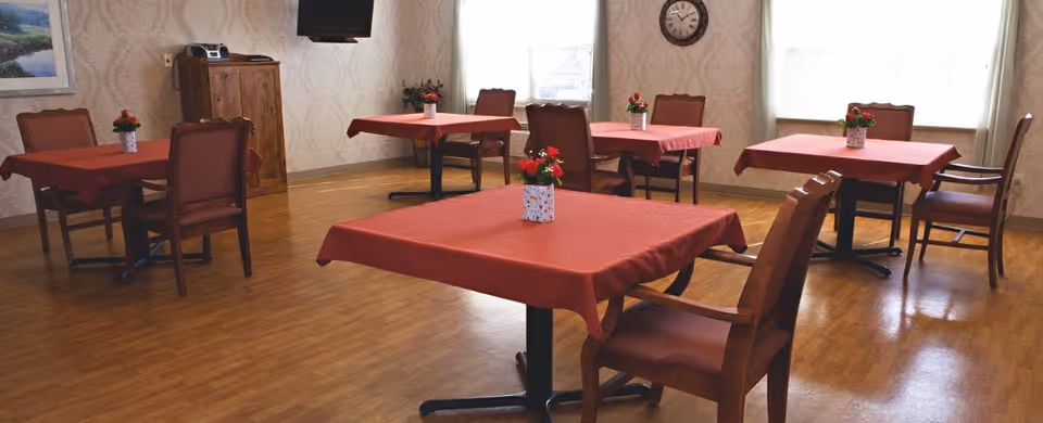 A dining room with several square tables covered with red tablecloths, each table having a small vase with red flowers. The room has wooden flooring, beige patterned wallpaper, a wall clock, a wooden cabinet with a stereo on top, and large windows letting in natural light.