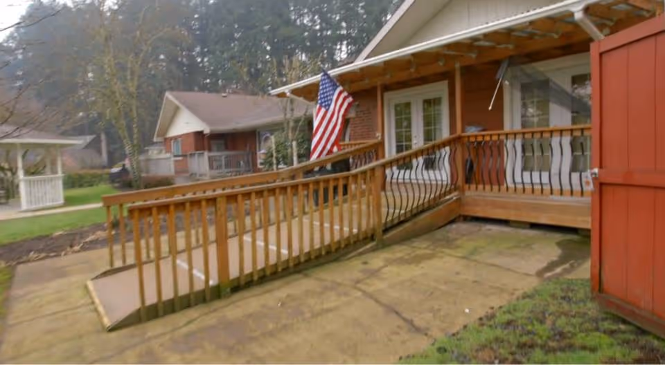 Outdoor view of a building entrance with a wooden wheelchair ramp and railing. An American flag is mounted near the door, and there is a red shed door open on the right side. The area is surrounded by trees and grass.