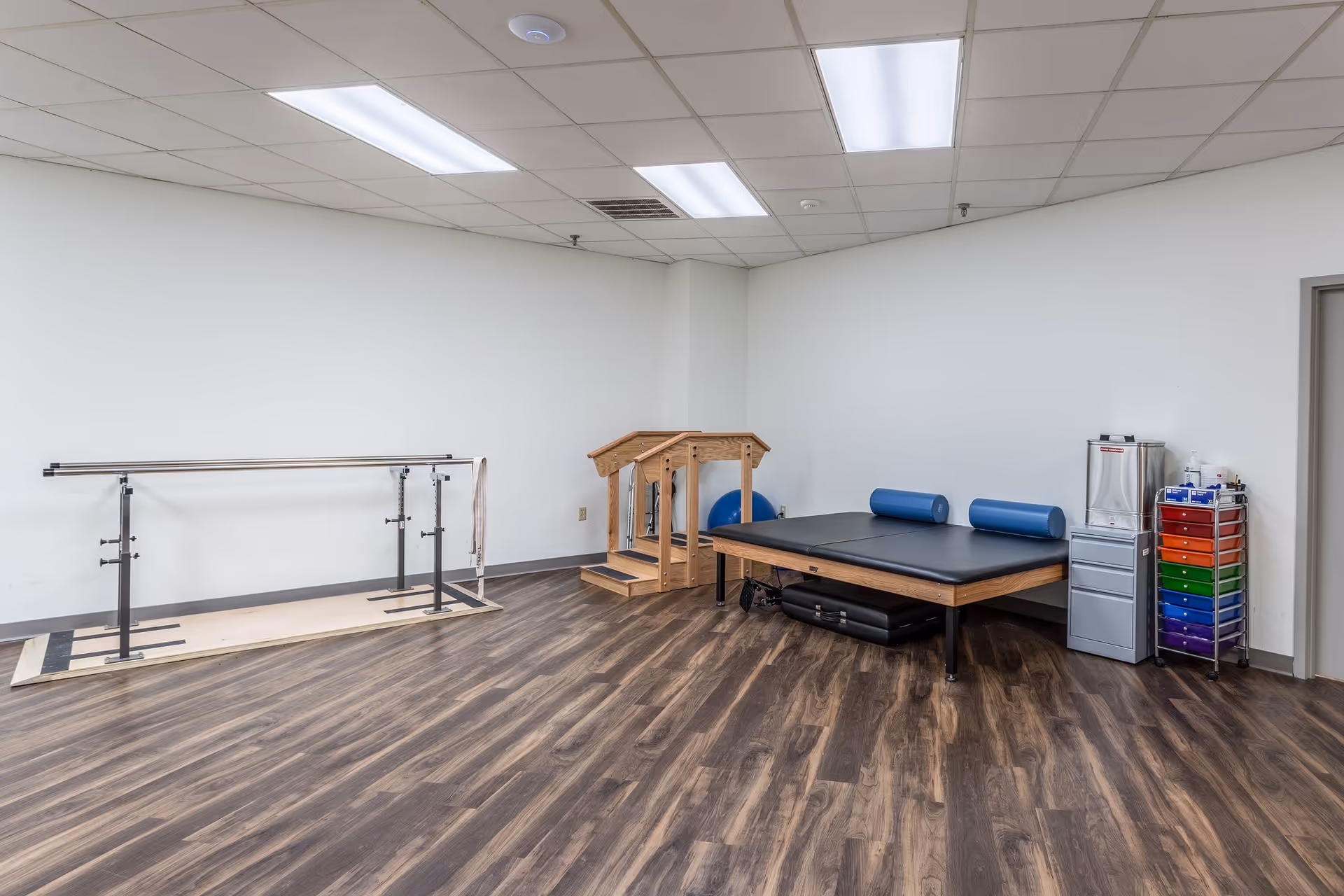 A physical therapy or rehabilitation room with wooden flooring, parallel bars for walking exercises, a padded therapy table with blue cylindrical cushions, a small wooden staircase, and a colorful storage cart with various supplies.