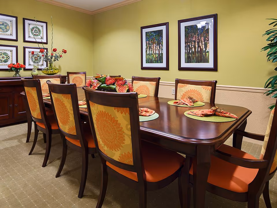 A dining room featuring a large wooden table surrounded by chairs with orange floral cushions, set with decorative napkins and placemats. The walls are painted a light green, adorned with framed artwork and a floral arrangement on the table.