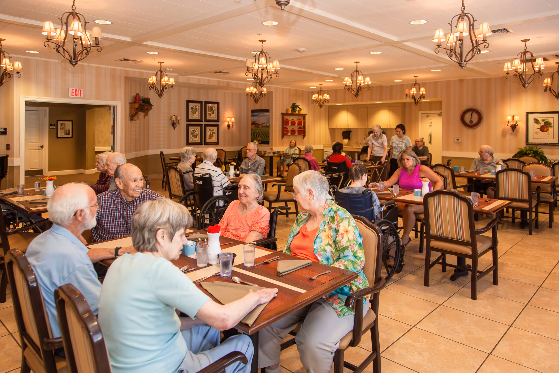 A dining room in an assisted living facility with elderly residents sitting at tables, some in wheelchairs, engaging in conversation and dining. The room is warmly lit with chandeliers and wall sconces, decorated with framed pictures and plants.