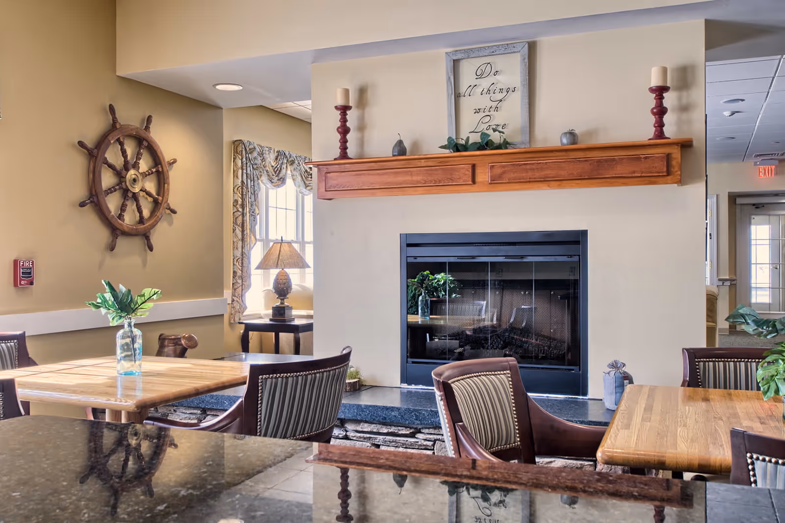 A cozy common area with wooden tables and chairs, a fireplace with a wooden mantel decorated with candles and a framed sign that reads 'Do all things with Love'. A ship's wheel hangs on the wall, and a window with patterned curtains lets in natural light. There are plants on the tables and a lamp on a side table near the window.