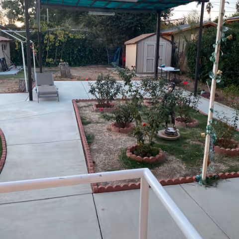 Outdoor garden area with a concrete pathway, small flower beds bordered with red bricks, a lounge chair, two storage sheds, and a covered patio with a green translucent roof. There are trees and bushes in the background.
