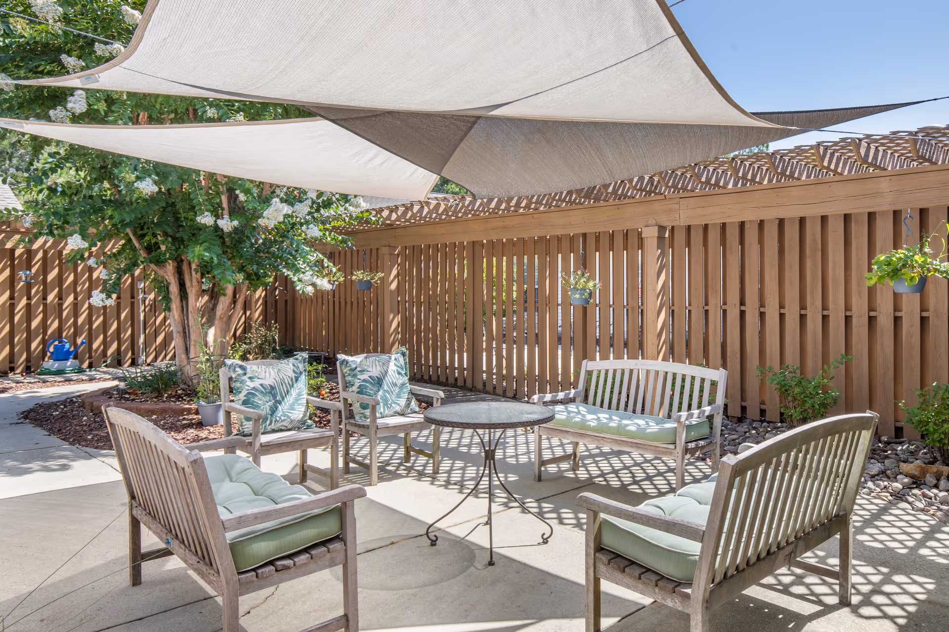 Shaded outdoor patio with wooden chairs and benches arranged around a round table next to a wooden fence and greenery.