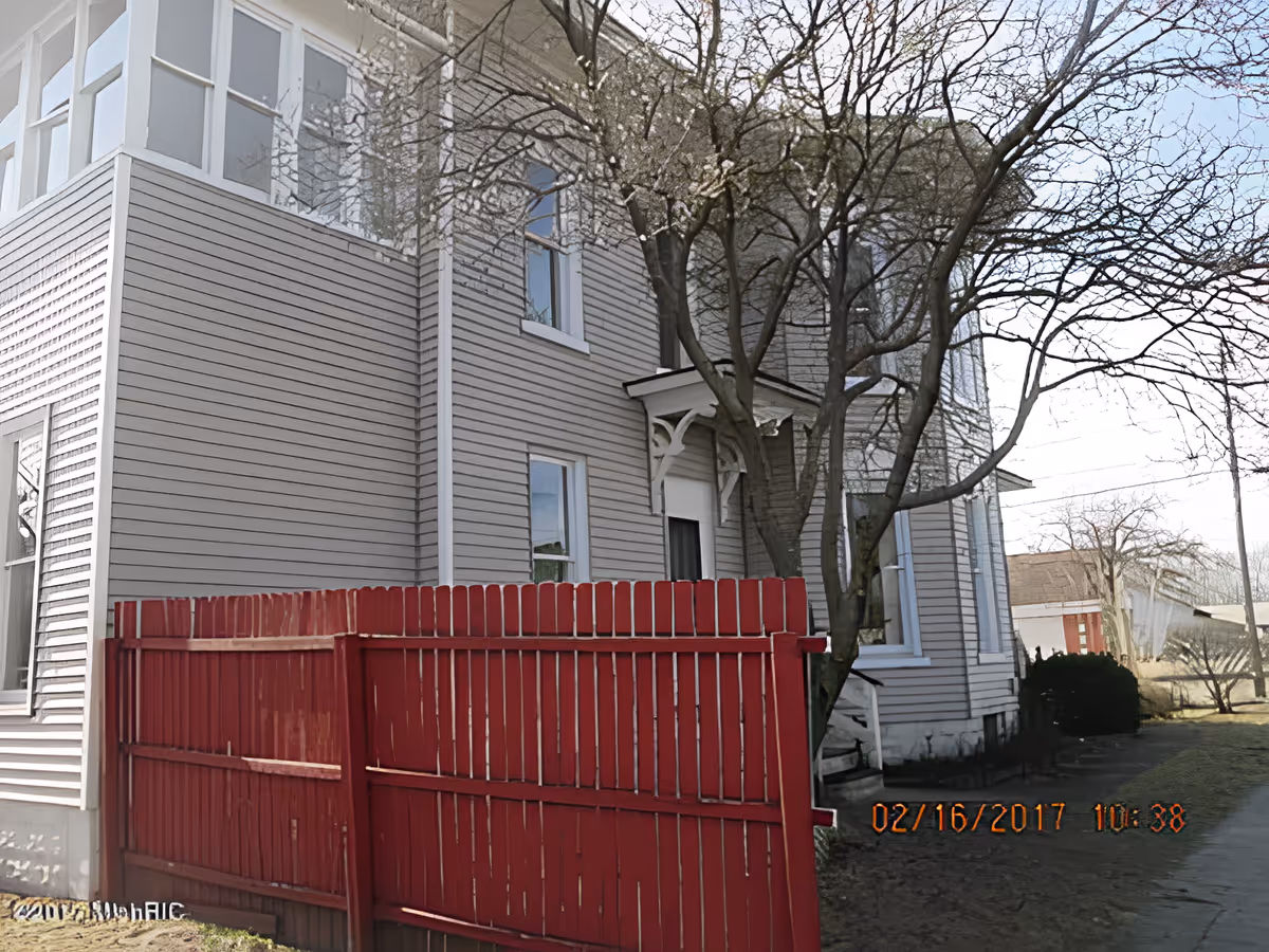 Gray two-story house with siding, a red wooden fence in front, and leafless trees by the entrance.