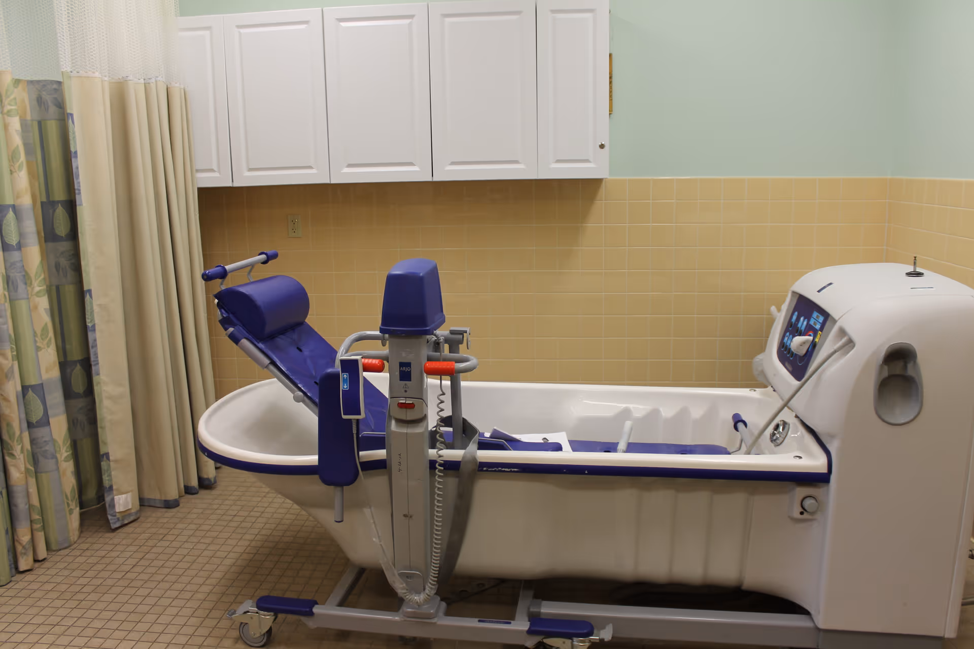 A specialized medical bathtub with a blue padded chair and mechanical lift system in a tiled room with beige and light green walls, white cabinets above, and patterned curtains on the left side.