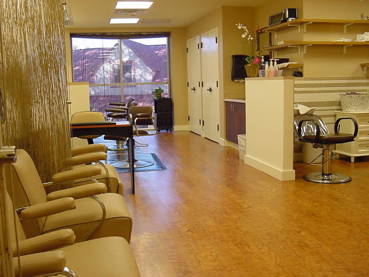 Interior salon area with multiple beige styling chairs, manicure tables, and a hair-washing station in a well-lit room.