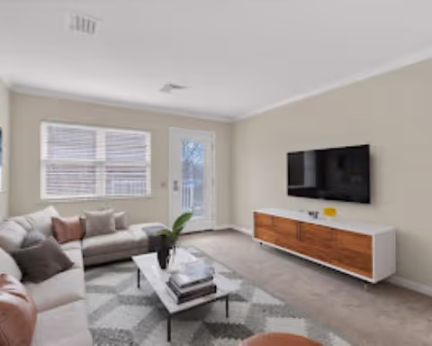 A bright living room with a large L-shaped beige sofa adorned with various cushions, a rectangular coffee table with books and a plant on it, a patterned area rug, a wall-mounted flat-screen TV, and a glass door leading outside. The walls are painted light beige and there is a window with blinds letting in natural light.