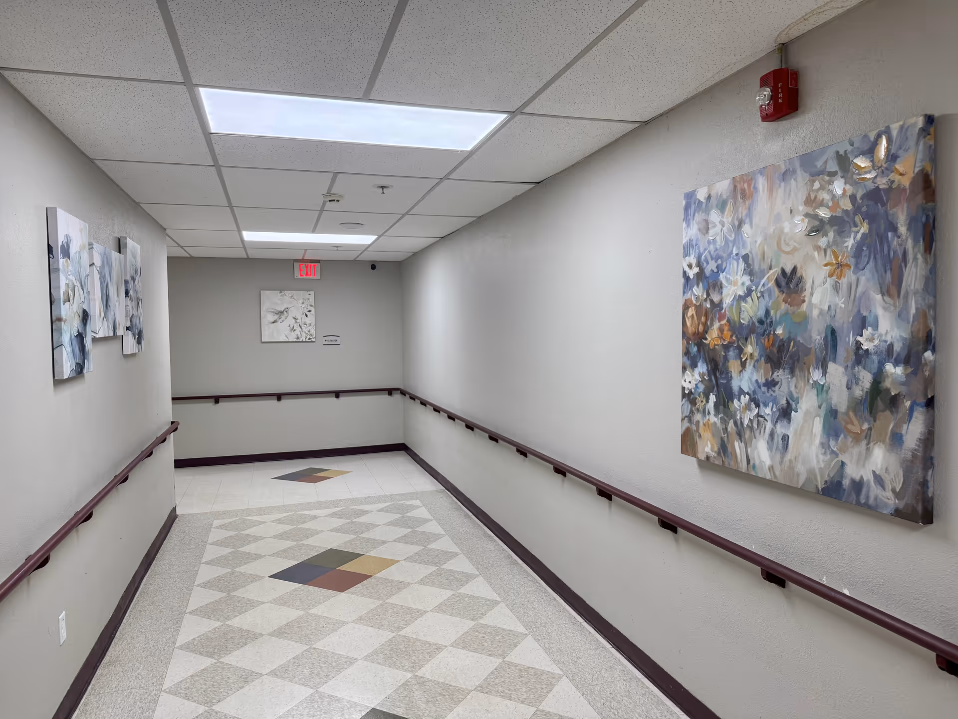 A well-lit hallway with handrails on both sides and decorative paintings on the walls. The floor has a patterned tile design with diamond shapes in various colors. There is an exit sign and a small framed picture at the end of the hallway.