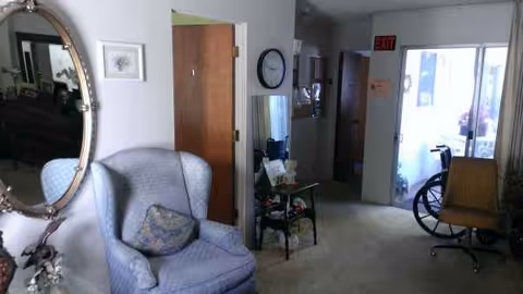 A cozy senior living lounge with chair, mirror, clock, and a hallway leading to an entrance.