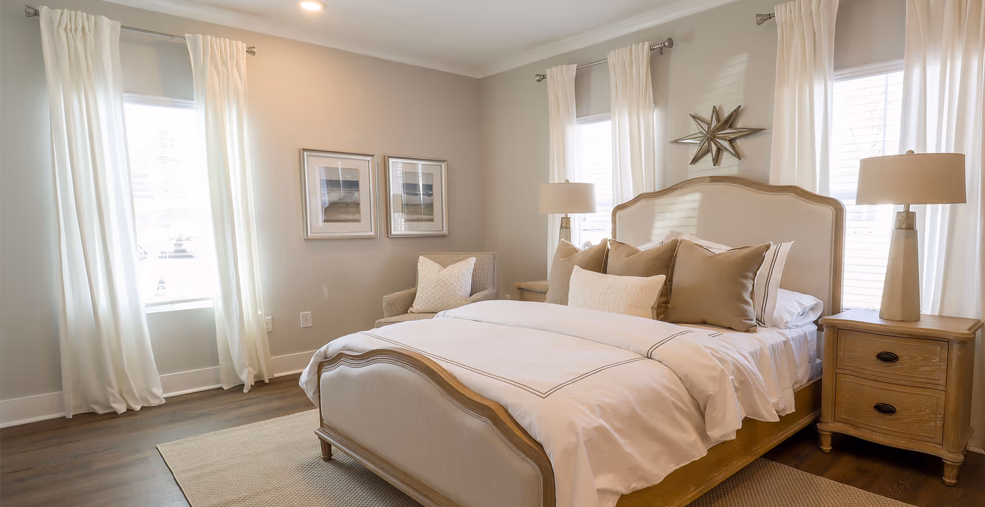 A bright and cozy bedroom with a large bed featuring beige and white bedding and multiple pillows. The room has two windows with white curtains, two wooden nightstands with matching lamps, a beige armchair with a pillow, two framed pictures on the wall, and a decorative star above the bed's headboard. The floor is wooden with a light-colored rug.