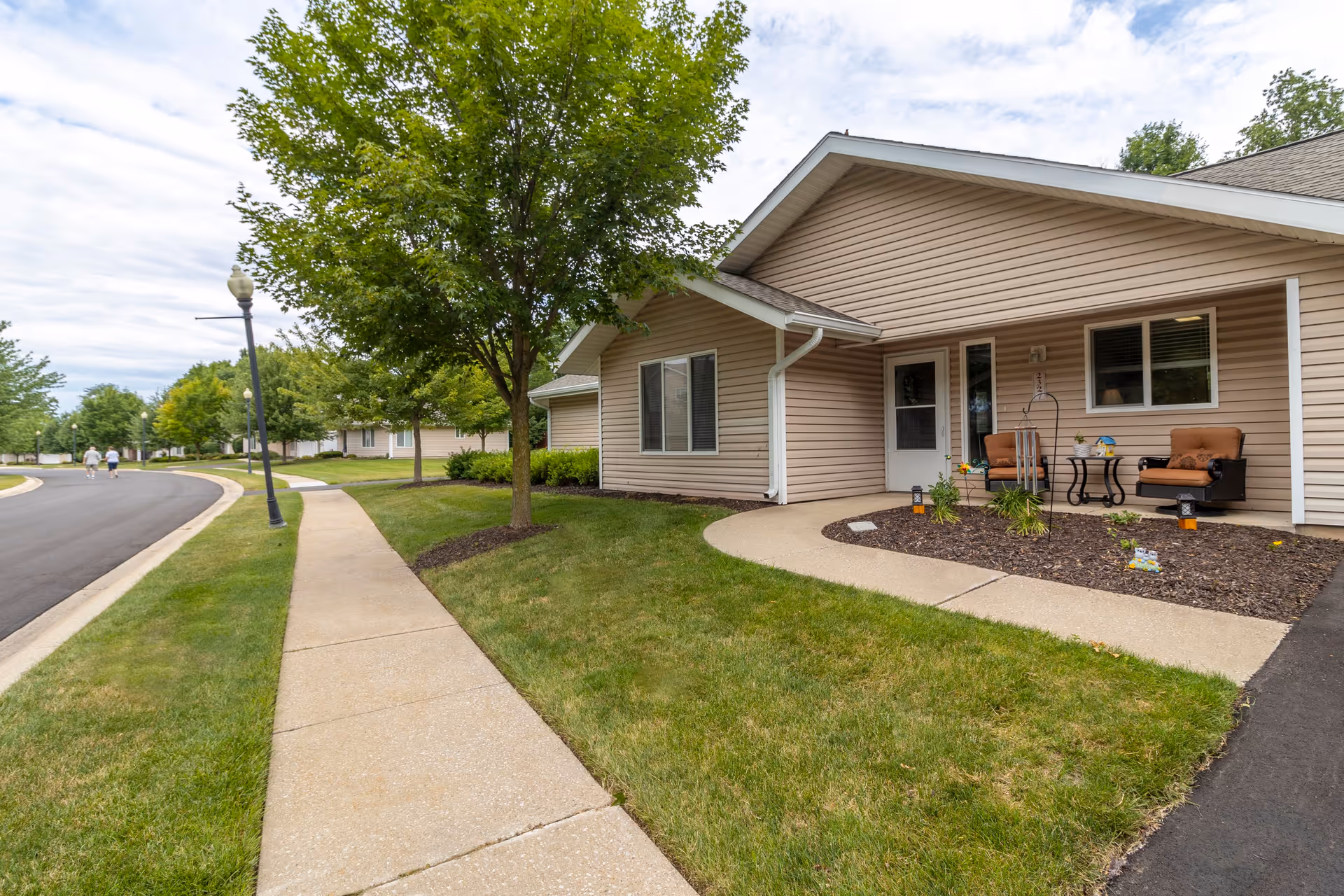 A sidewalk runs alongside a beige single-story building with a small porch area furnished with two cushioned chairs and a small table. The building is surrounded by green grass, trees, and landscaped mulch beds. In the background, there are more similar buildings and a street with two people walking. The sky is partly cloudy.