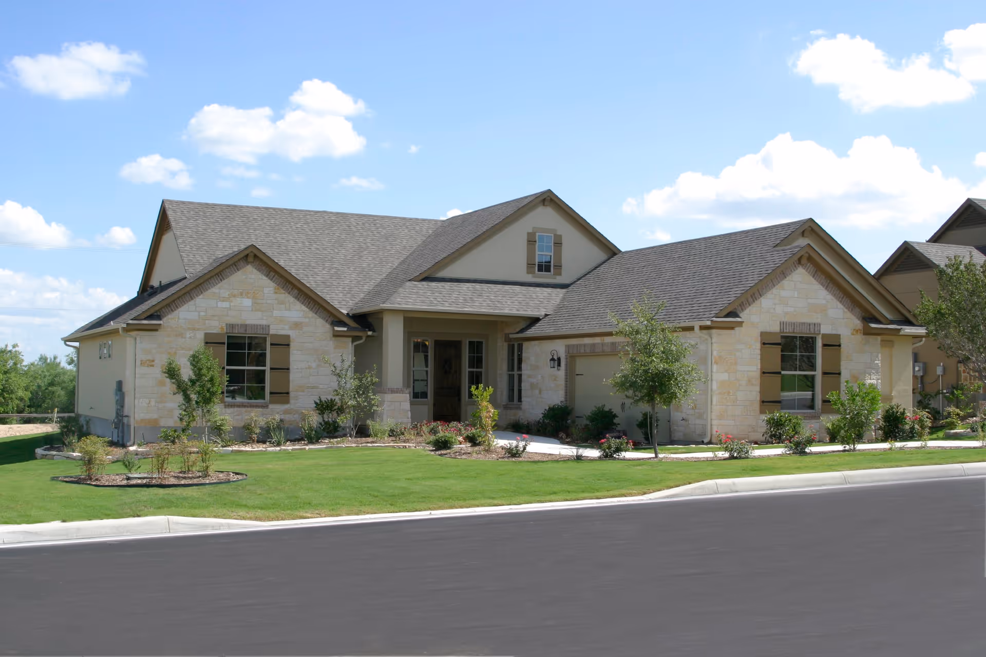 Single-story residential building with stone and beige siding, a gray shingled roof, and a small front porch. The front yard is landscaped with green grass, small trees, and bushes. The sky is clear with a few clouds.
