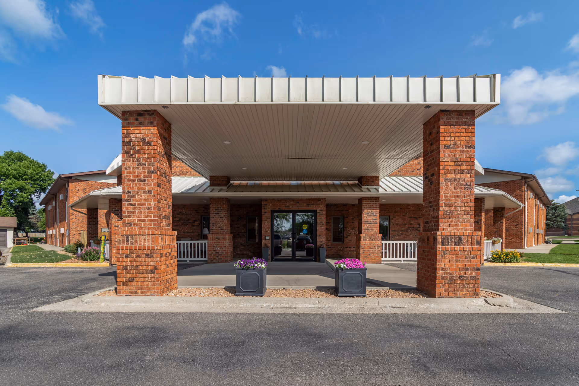 Front entrance of a senior living facility with a covered drop-off area supported by brick columns, two planters with purple and pink flowers, and a glass door entrance under a clear blue sky.