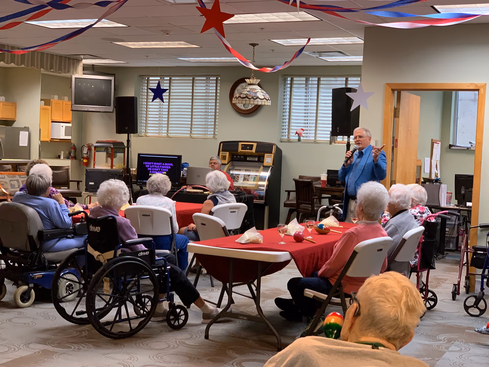 A group of elderly people seated around tables with red tablecloths in a common room, watching a man in a blue jacket holding a microphone and speaking. The room is decorated with red, white, and blue streamers and stars. There is a TV screen displaying karaoke lyrics, a jukebox, and a kitchen area in the background.