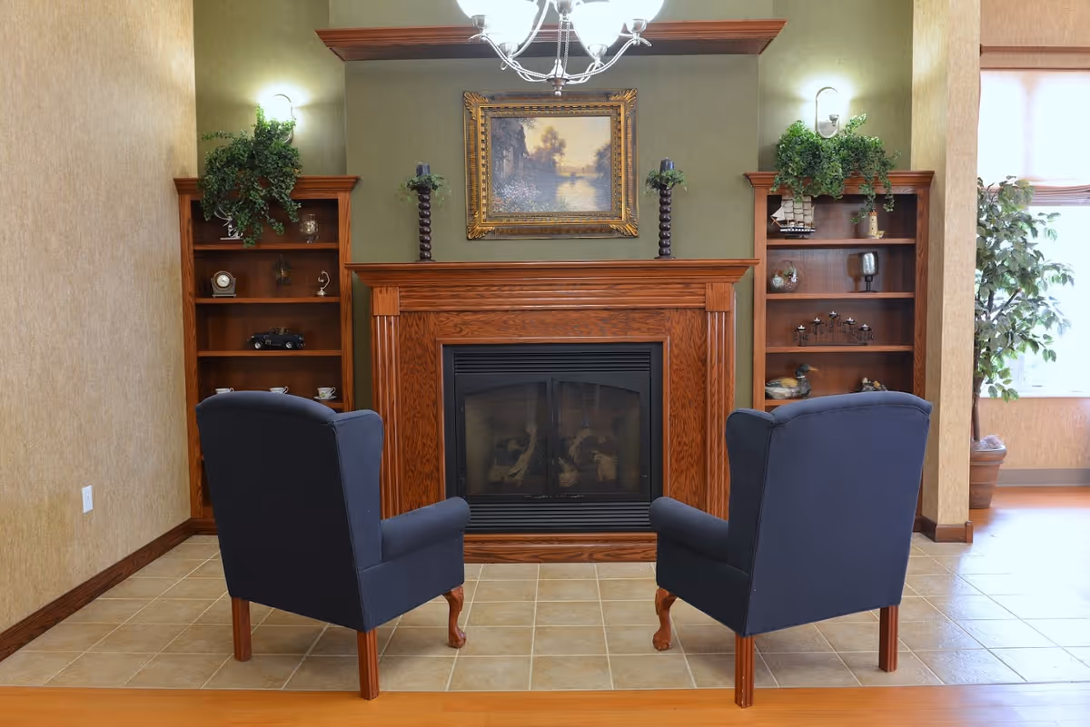 A cozy living room area featuring two dark blue armchairs facing a wooden fireplace with a framed painting above it. On either side of the fireplace are wooden built-in shelves decorated with various ornaments and plants. The room has tiled flooring and warm lighting.