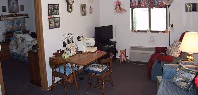 Interior view of a senior living facility room showing a small dining table with two wooden chairs, a sewing machine on the table, a television on a stand, a window with floral curtains, a blue armchair with a decorative pillow, a red recliner, and a bed partially visible in an adjacent room.