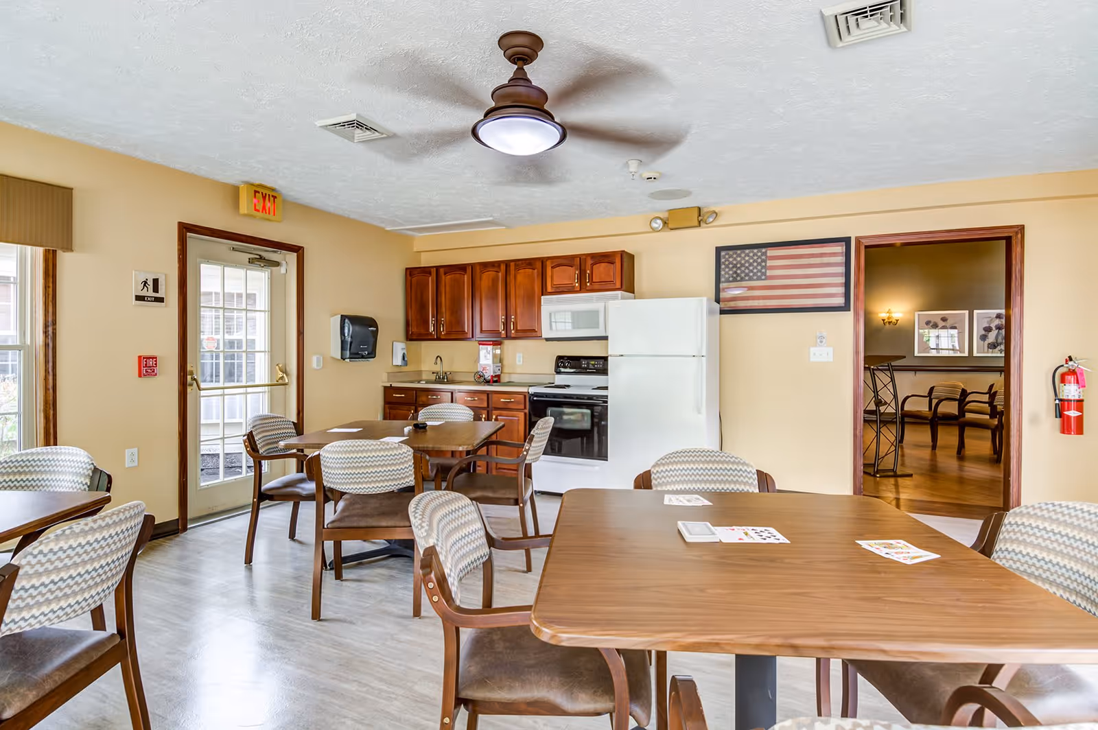 A communal dining area with several wooden tables and chairs featuring patterned cushions. The room includes a small kitchen area with wooden cabinets, a white refrigerator, stove, and microwave. There is an American flag framed on the wall, a ceiling fan with a light, and a door leading outside with an exit sign above it. A fire extinguisher is mounted on the wall near an open doorway leading to another room with additional seating and framed artwork.