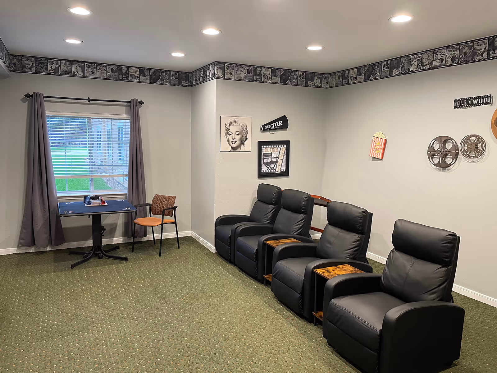 Small lounge/media room with four black recliners in a row, movie-themed wall decor, and a window with a table and chairs.
