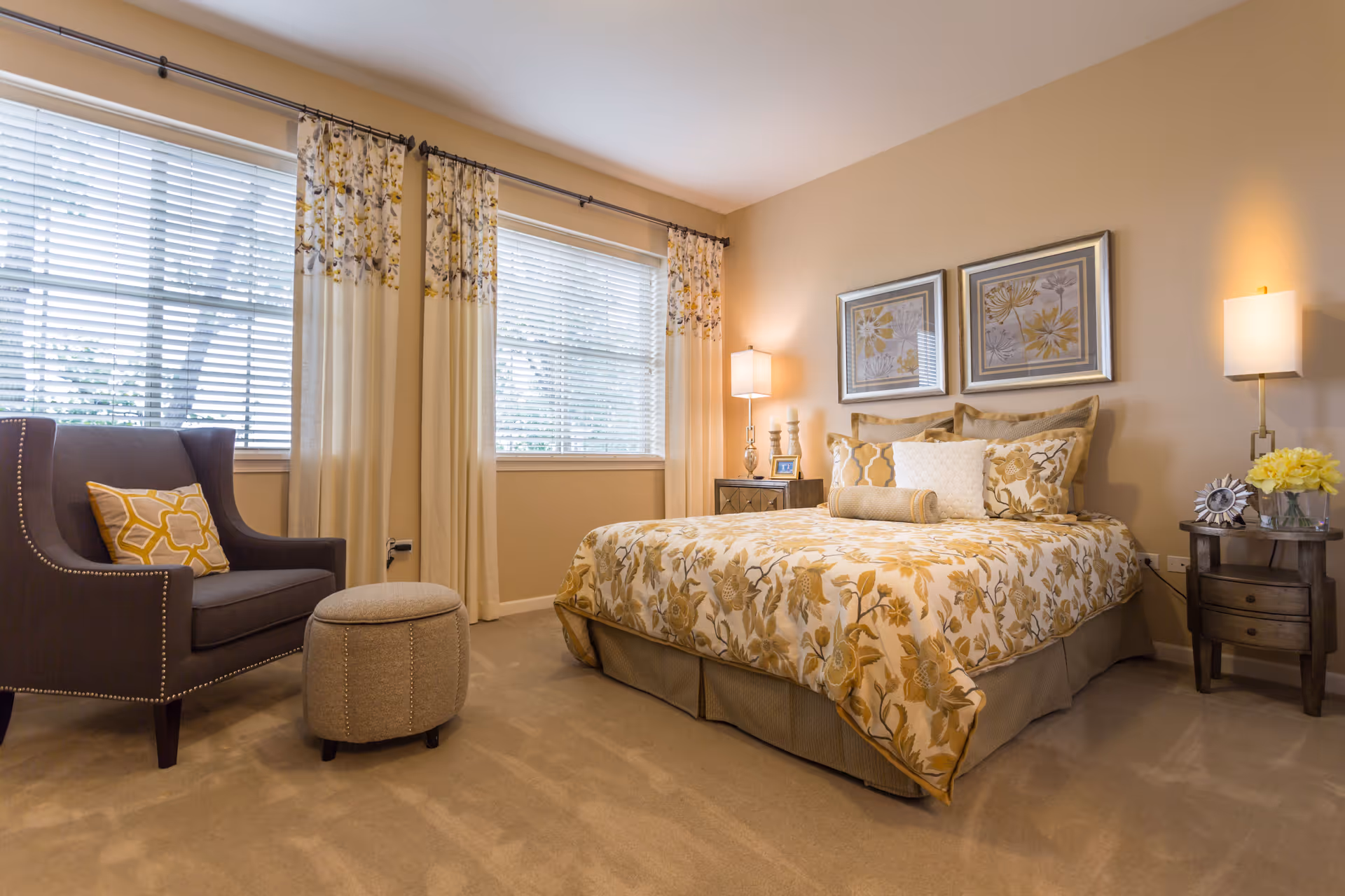 A cozy bedroom with a large bed covered in floral patterned bedding in shades of yellow and beige. There are two framed floral artworks hanging above the bed. On either side of the bed are wooden nightstands with lamps, decorative items, and a vase of yellow flowers. A dark upholstered armchair with a yellow patterned pillow and a small round ottoman are positioned near two large windows with white blinds and cream-colored curtains with floral accents.