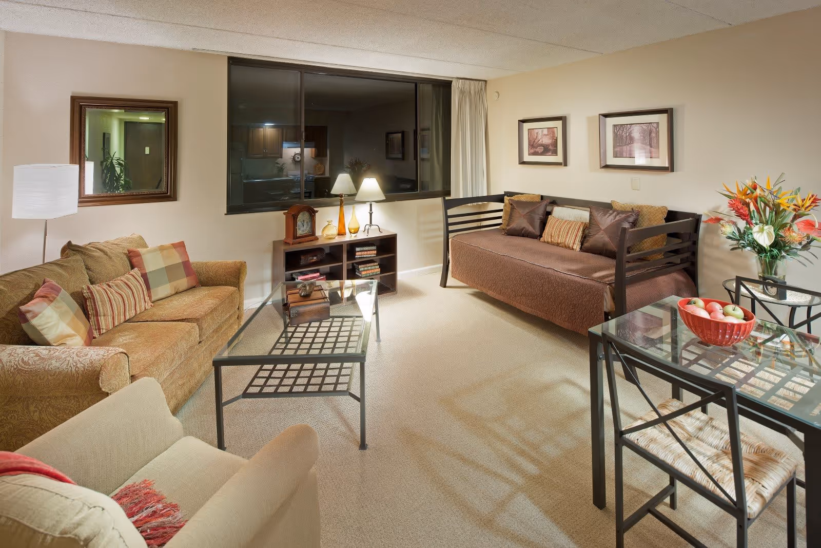A cozy living room in a senior living facility with a beige armchair, a patterned sofa with multiple cushions, a daybed with pillows, a glass coffee table, a small wooden shelf with books and decorative items, a floor lamp, a table lamp, framed pictures on the wall, and a glass dining table with chairs. A bowl of apples and a vase with colorful flowers are on the dining table.