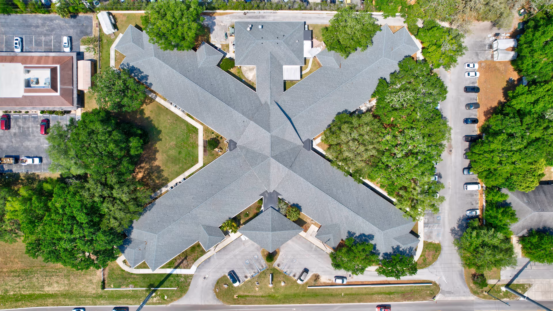 Aerial view of the Gardens of Westbrooke senior living facility showing a large building with a unique X-shaped roof surrounded by trees, parking lots, and roads.